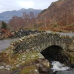 Scenic view of Ashness Bridge in the Lake District, with stone arch crossing a rocky stream and mountains visible in the background.